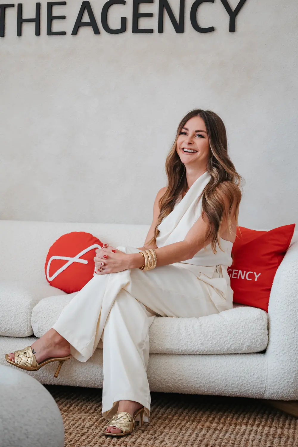 Agent seated beneath a The Agency sign in a cream jumpsuit